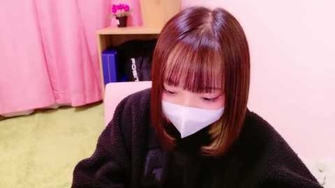 Video of an Asian woman with shoulder-length brown hair, wearing a black robe and white medical mask, sitting on a green carpet in a pink room with a wooden shelf and pink curtains.