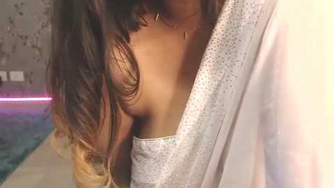 Media: Video of a partially visible, slender woman with long, wavy, ombre hair, wearing a sheer, glittery dress and a delicate necklace, in a dimly lit room with a blue pool in the background.