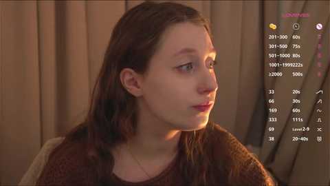 Video of a young woman with fair skin and brown hair, wearing a brown knit sweater, looking pensive against a beige curtain backdrop.