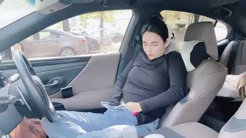 Media: Video of a pregnant woman in a car seat, adjusting the seatbelt, wearing a dark turtleneck and jeans, with a blurred background of parked cars.