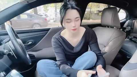 Media: A video of a young woman with fair skin and dark hair tied back, sitting cross-legged in the driver's seat of a car, wearing a black top and blue jeans, looking at a phone.