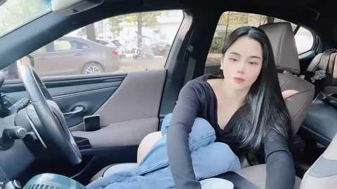 Media: A video of an Asian woman with long black hair, wearing a black top and jeans, seated in a car, holding a child on her lap, with a parked car visible outside.