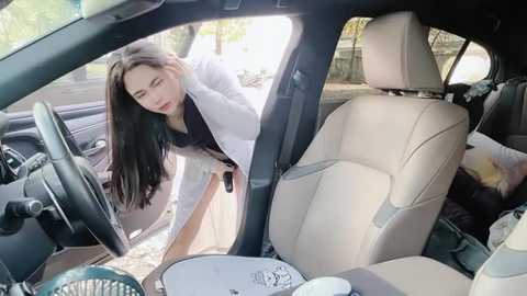 Media: Video of a young Asian woman with long black hair, dressed in a white lab coat, bending over to check the car's undercarriage. The car interior features beige leather seats and a cluttered dashboard.