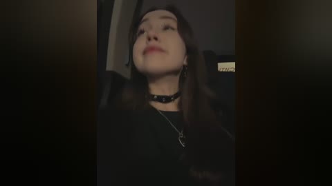 Media: Video of a young woman with long brown hair, wearing a black choker and top, looking up with a neutral expression.