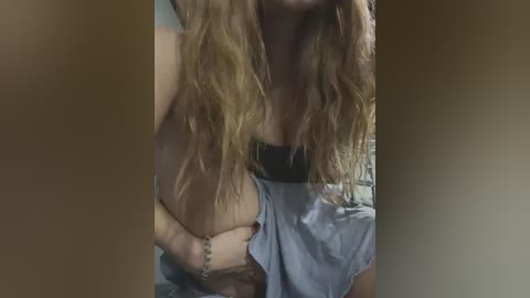 Media: Video of a woman with long, wavy blonde hair, wearing a blue blouse and black bra, clutching her left breast, blurred background.