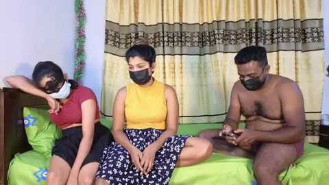 Media: Video of three South Asian individuals in a bedroom: two women wearing masks, one man with a beard, all seated on a green bed with patterned sheets, against a backdrop of yellow and black curtains.