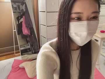 Media: A video of an Asian woman with long black hair, wearing a white face mask, beige sweater, and sitting on a pink blanket in a cluttered bedroom with wooden floor, closed closet, and white shelves.