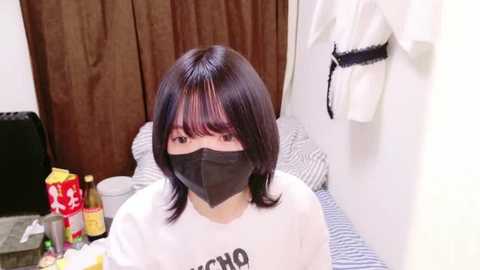 Media: Video of an Asian woman with shoulder-length dark hair, wearing a black face mask, white t-shirt, and sitting at a cluttered desk. Background includes a brown curtain, white clothing on a rack, and a messy room.