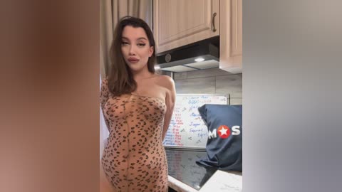 Media: A video of a young woman with fair skin and medium-length brown hair, wearing a sheer, leopard-print bodysuit, standing in a modern kitchen with light cabinets, a stove, and a whiteboard.