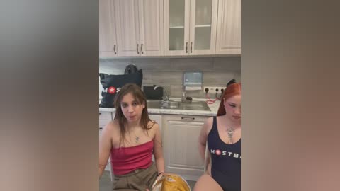 Media: Video of two women in a modern kitchen; one with brown hair, red top, and beige shorts, the other with red hair, blue top, and visible tattoo, both seated at a counter.