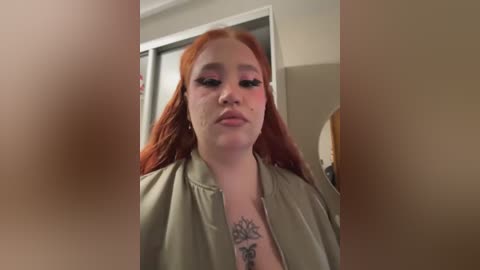 Media: Video of a young woman with long, vibrant red hair, wearing a light green jacket. She has a tattoo on her chest and stands in a dimly lit room with beige walls and a mirror.