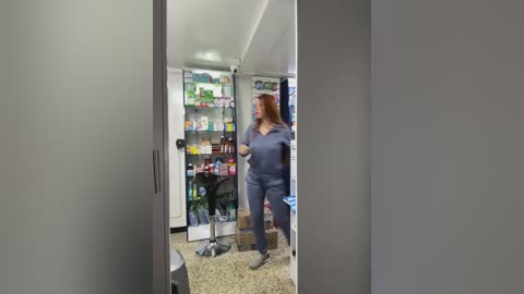 Media: A video of a woman in blue scrubs standing in a kitchen, opening a refrigerator filled with various food items and beverages. The background includes white walls and a speckled beige floor.