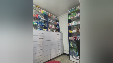 Video of a well-organized pharmacy with white cabinets, shelves, and drawers filled with colorful medicines and vitamins. The shelves are neatly stacked, and the floor is covered in speckled tiles.