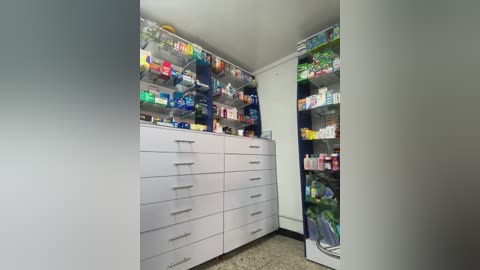 Media: A video of a well-organized pharmacy room with white cabinets and drawers on the left, shelves filled with colorful medicine bottles on the right, and a beige tiled floor.