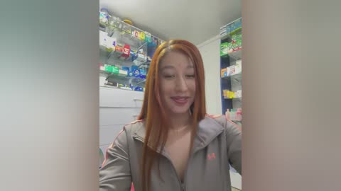 Media: Video of a smiling Asian woman with long red hair, wearing a gray hoodie, standing in a brightly lit pharmacy with shelves filled with colorful medicine bottles.