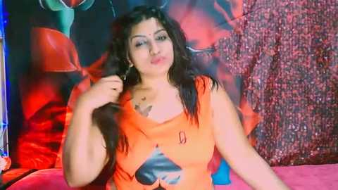 Media: Video of a dark-skinned woman with curly hair, wearing an orange top with a black heart design, posing against a colorful, abstract backdrop.