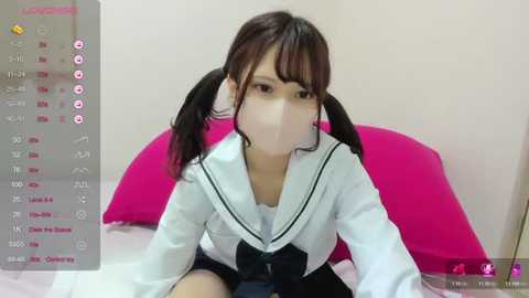 Media: A video of an East Asian woman with pigtails, wearing a white sailor school uniform, sitting on a bed with a pink pillow, indoors.