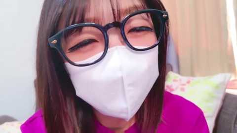 Media: Video of an Asian woman with straight, shoulder-length brown hair, wearing large black-framed glasses and a white surgical mask, standing indoors in a brightly lit room with a green floral pillow.