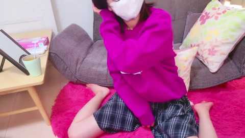 Media: Video of an Asian woman with short black hair, wearing a bright pink sweater and plaid shorts, sitting on a red rug in a living room with a grey couch, floral pillows, and wooden furniture.