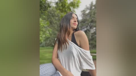 Media: Video of a young woman with long brown hair, wearing a white off-shoulder top, standing in a lush green park, looking upward.