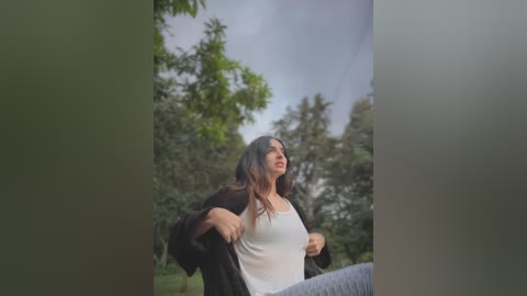 Media: Video of a young woman with long brown hair, wearing a white tank top and black jacket, standing outdoors in a lush, green forest.