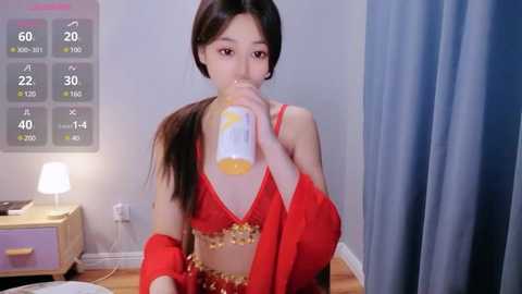 Media: A video of an Asian woman with long dark hair, wearing a red bra and gold accessories, drinking juice in a modern, minimalistic room with blue curtains.