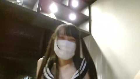 Media: A video of a woman wearing a surgical mask and a black sailor-style school uniform with white accents, standing in a dimly lit room with a black shelving unit and overhead lighting.