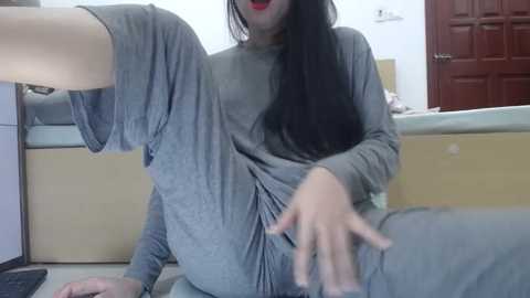 Media: Video of an Asian woman with long black hair, wearing a loose gray t-shirt, sitting on a kitchen counter, legs spread, one hand on her thigh, mouth open, in a casual, possibly provocative pose.