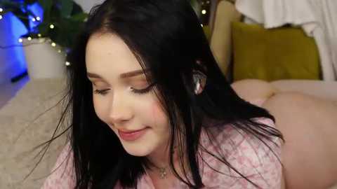 Video of a young woman with long black hair, fair skin, and pink floral robe, lying on a beige carpet in a cozy room with potted plants, yellow pillows, and fairy lights.