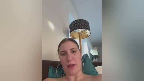 Media: Video of a fair-skinned, brown-haired woman with large hoop earrings, seated on a bed with teal pillows, looking relaxed. Background includes a lamp with a geometric shade and a blurred, modern living room.