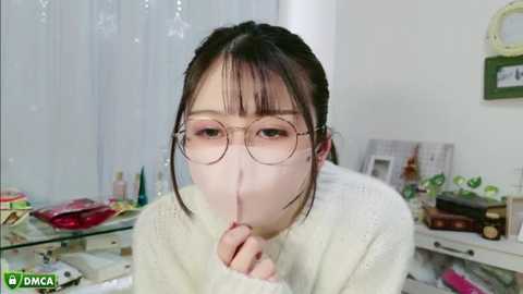 Media: A video of an Asian woman with fair skin, dark hair, and round glasses, wearing a white sweater, covering her mouth with her fingers, in a cluttered, softly lit room with a white curtain, various personal items, and a small framed picture.