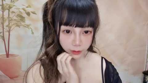 Media: Video of a young Asian woman with long black hair, fair skin, and a neutral expression. She's wearing a black strapless top. Background features a potted plant and a soft, beige wall.