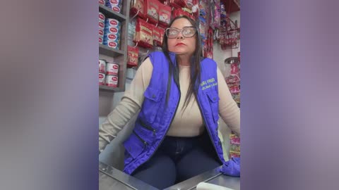 Media: Video of a Latina woman in a convenience store, wearing a blue vest over a beige shirt and dark jeans, glasses, and a serious expression. Shelves with snacks and drinks in the background.