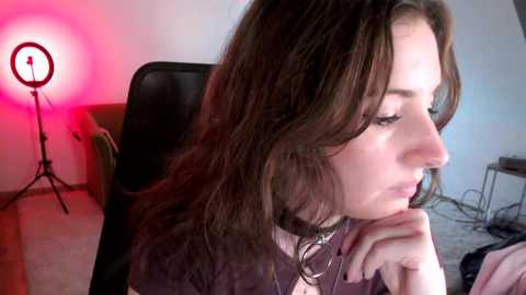 Media: Video of a young woman with wavy brown hair, fair skin, and a contemplative expression, sitting in a black chair under a pink light.