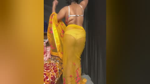 Media: Video of a woman in a yellow sari with red floral embroidery, revealing her white bra and backside. She stands in a dimly lit room, arms raised.