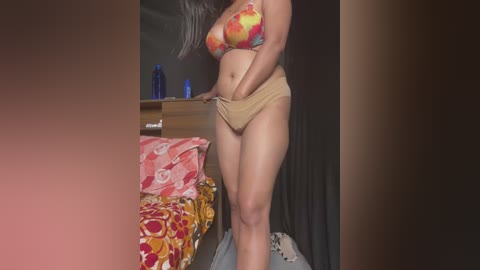 Media: A video of a curvy woman with long dark hair in a colorful bikini, adjusting beige shorts in a dimly-lit bedroom with floral-patterned bedding and a blue water bottle.