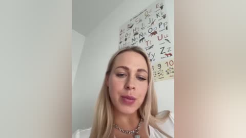 Media: Video of a blonde woman with fair skin, wearing a white top and silver necklace, posing with a pouty expression in a room with a white wall and science-themed poster.