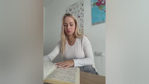 Media: Video of a blonde woman in a white long-sleeve shirt, studying an open notebook on a desk, in a minimalist, white-walled classroom with a colorful world map and educational posters.