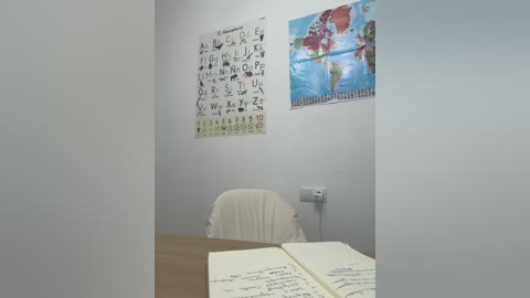 Media: Video of a small, sparsely furnished classroom with light gray walls, featuring a white chair, a wooden table with an open notebook, and educational posters of a world map and a phonics chart.