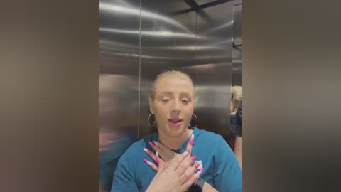 Media: Video of a blonde woman in teal scrubs with pink nails, hands clasped in prayer, in a metallic elevator with blurred background.