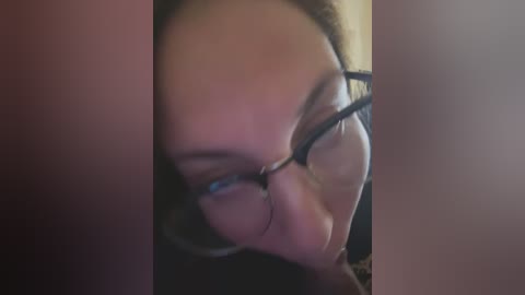 Media: A close-up video of a young woman with light skin, wearing black-framed glasses and a dark jacket, focusing intently on a screen. The background is blurry, emphasizing her face.