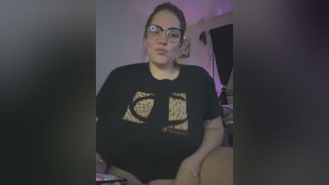 Media: Video of a young woman with glasses, light skin, and dark hair tied back, wearing a black graphic tee featuring a leopard print design, sitting in a dimly lit room with purple lighting.