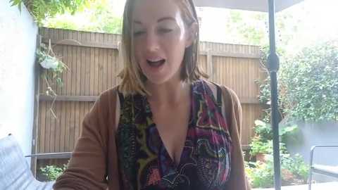 Media: Video of a fair-skinned woman with shoulder-length light brown hair, wearing a colorful, patterned blouse, laughing outdoors under a patio umbrella, with a wooden fence and green plants in the background.