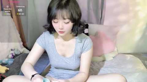 Media: A young East Asian woman with shoulder-length black hair, wearing a light gray t-shirt, sits cross-legged on a bed, playing a video game. The background features pastel curtains and scattered toys.