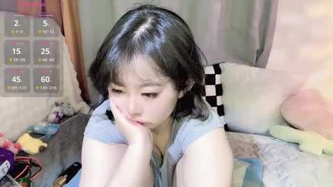 Media: Video of an Asian woman with short black hair, light skin, and small breasts, lying on a bed with stuffed animals, wearing a light grey t-shirt.