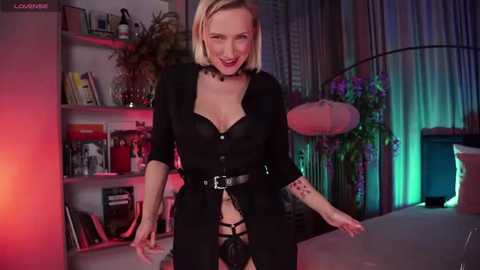 Media: A video of a blonde, fair-skinned woman in a black, lace-up corset with a visible penis, standing in a dimly lit, modern room with shelves, flowers, and a TV.