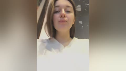 Media: Video of a young woman with light skin and straight brown hair, wearing a white V-neck shirt, standing indoors with a blurred background.