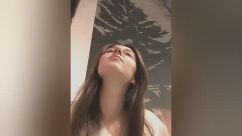 Media: A video of a young woman with long, straight brown hair, wearing a white tank top, looking upward with a serene expression. The background features a large, dark silhouette of a palm tree on a light-colored wall.