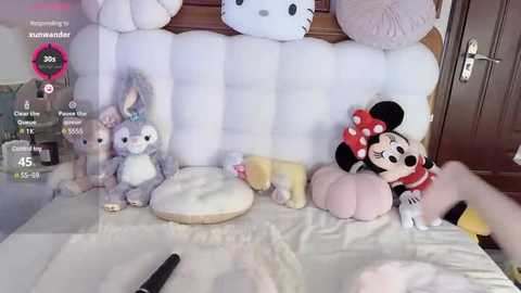 Media: Video of a plush Minnie Mouse sitting on a white bed, surrounded by stuffed animals including a Hello Kitty and two bunny toys.