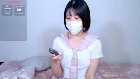 A video of an East Asian woman with short black hair, wearing a white mask, white top, and pink plaid scarf, holding a smartphone in a bedroom with pink bedspread.